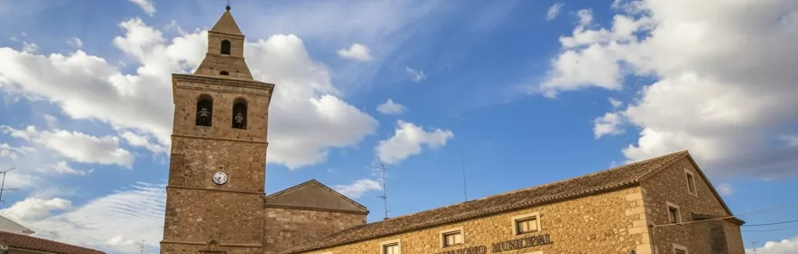 Plaza con torre de piedra y edificio histórico bajo cielo azul con nubes.