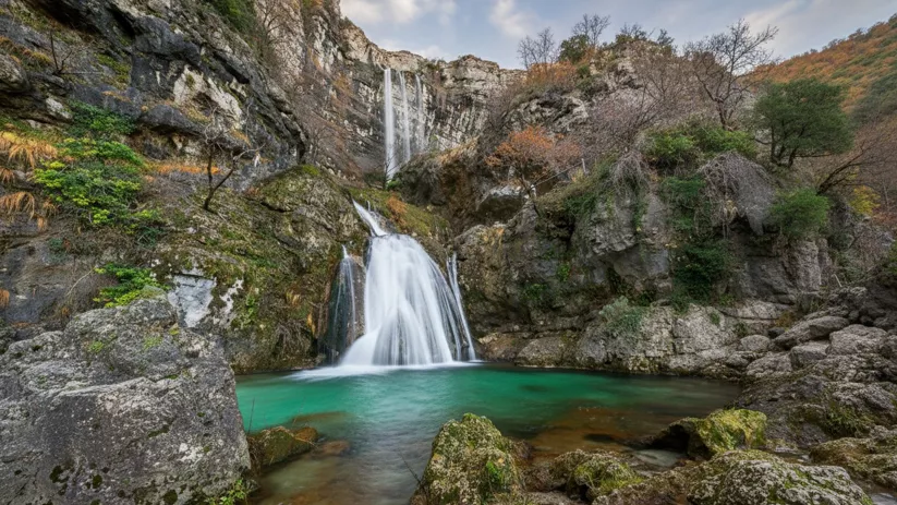 Cascada doble hacia una poza verde turquesa, rodeada de grandes rocas y vegetación.