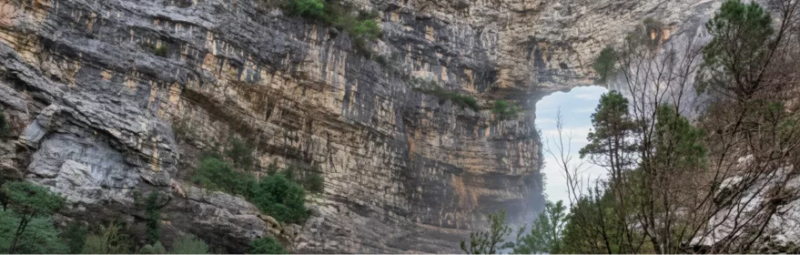Pared de roca con gran abertura natural, rodeada de árboles y matorral.