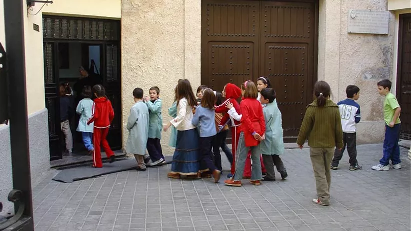 Grupo de niños y niñas entrando en fila a un edificio histórico por una puerta lateral.