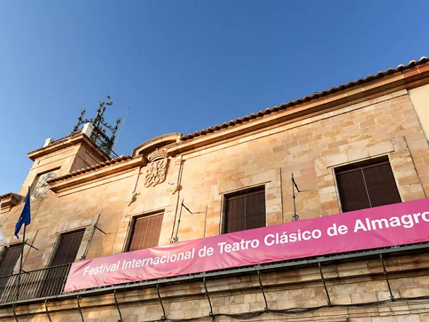 Fachada de edificio histórico de piedra con balcón y pancarta cultural, bajo cielo azul despejado.