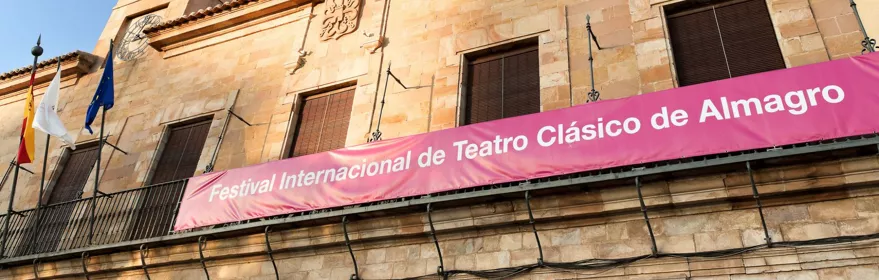 Fachada de edificio histórico de piedra con balcón y pancarta cultural, bajo cielo azul despejado.