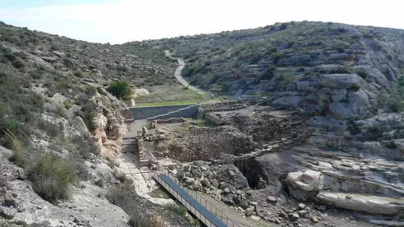 Valle rocoso con pasarela peatonal, restos arqueológicos y laderas escarpadas.