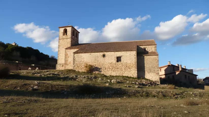 Vista lateral de iglesia de piedra en un campo abierto, con casas cercanas.