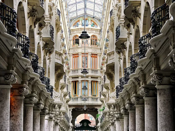 Galería interior simétrica con columnas de piedra, balcones de hierro y techo acristalado.
