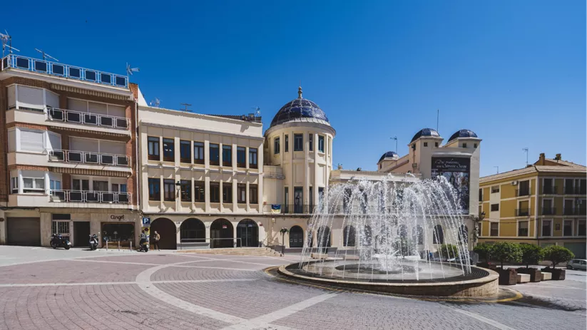 Fuente en funcionamiento en una plaza amplia con edificio histórico de cúpulas azules al fondo.