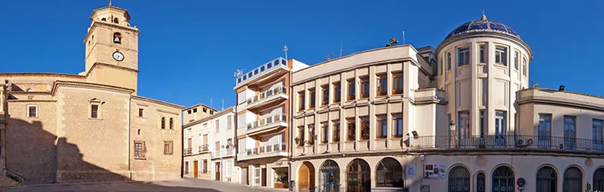 Vista panorámica de una plaza amplia con edificio institucional de cúpula azul y torre de piedra a un lado.