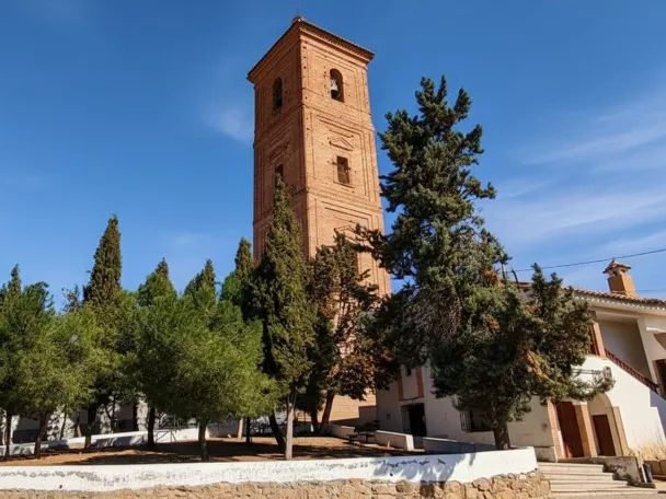 Torre de ladrillo rodeada de pinos y edificios encalados en día soleado.