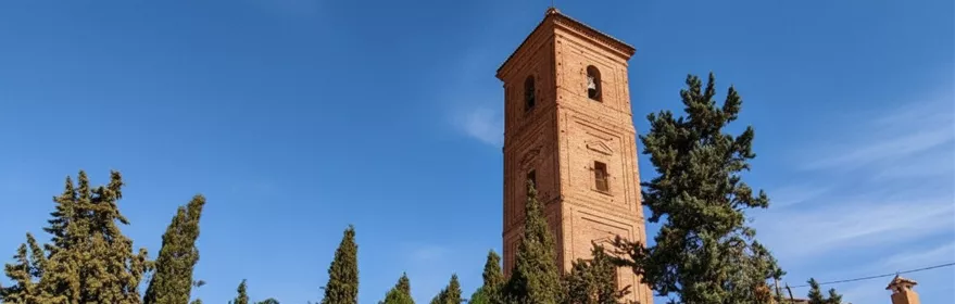 Torre de ladrillo rodeada de pinos y edificios encalados en día soleado.