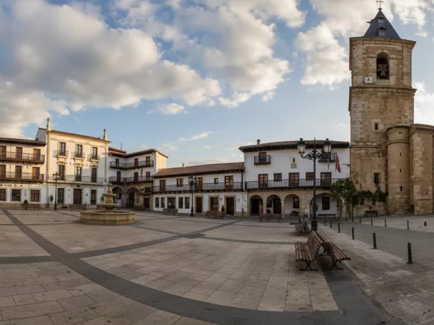 Plaza amplia con fuente central, edificios con balcones y torre de iglesia al fondo.