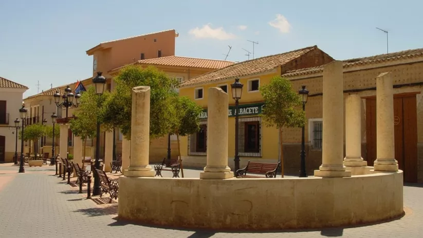 Plaza de Pozo Cañada con columnas de piedra, bancos y edificios tradicionales.