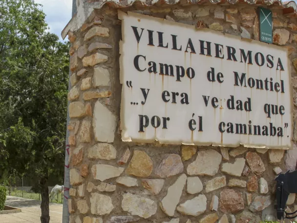 Señal de bienvenida de piedra junto a una avenida arbolada y una iglesia al fondo.