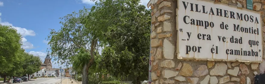 Señal de bienvenida de piedra junto a una avenida arbolada y una iglesia al fondo.