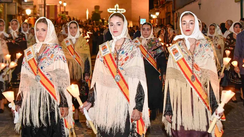 Mujeres con mantilla y banda portando velas en una procesión nocturna.