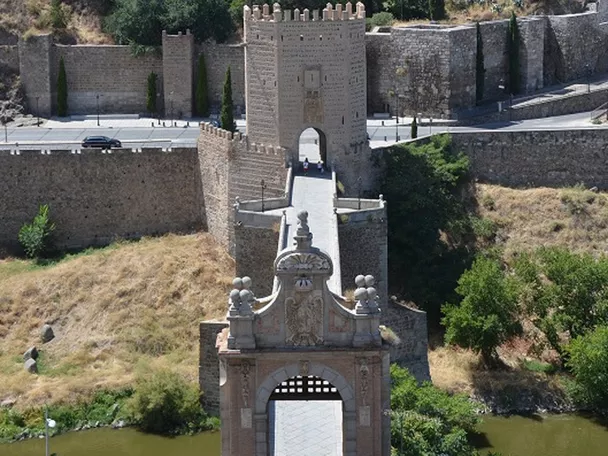 Puente de piedra con torre fortificada y murallas sobre un río, con ciudad histórica al fondo.