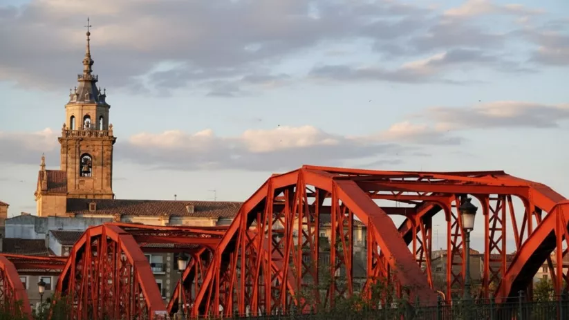 Estructura de puente rojo con una torre de iglesia al fondo.