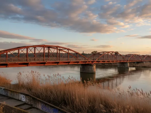 Puente de hierro rojo sobre un río al atardecer, con juncos en primer plano.