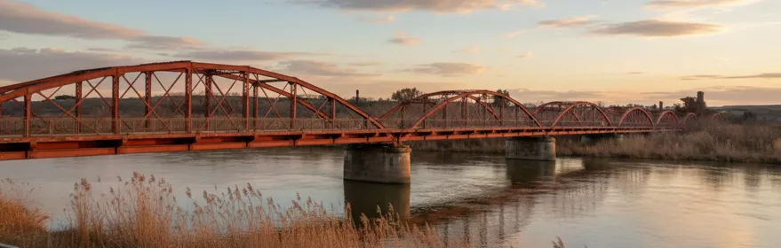 Puente de hierro rojo sobre un río al atardecer, con juncos en primer plano.