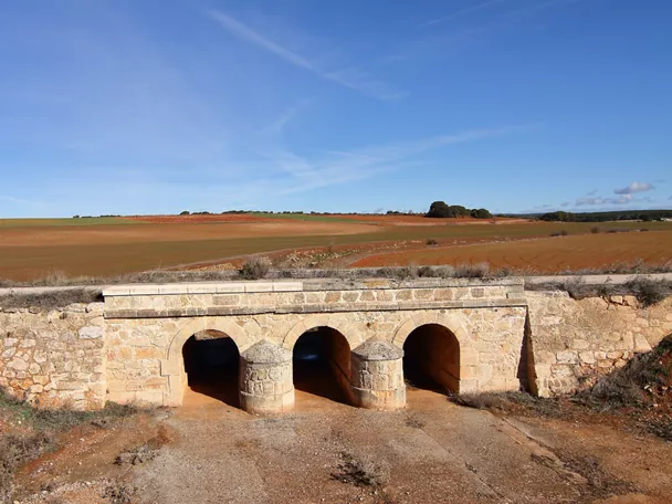 Puente de piedra con tres arcos sobre un camino rural.