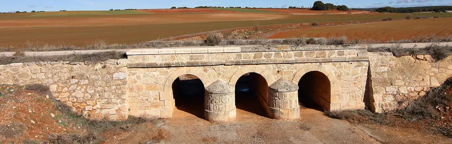 Puente de piedra con tres arcos sobre un camino rural.