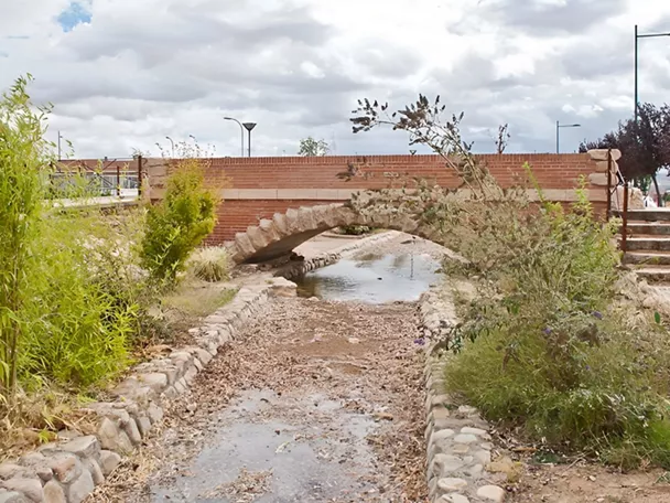 Arroyo canalizado con pequeño puente de piedra y vegetación.