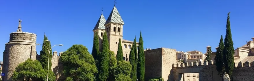 Muralla histórica con torre cuadrada y cipreses junto a la carretera.