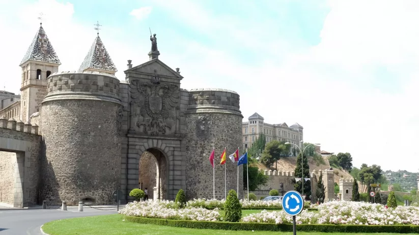 Acceso fortificado con escudo tallado y banderas frente a glorieta.