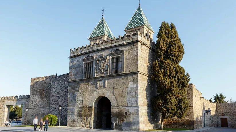 Puerta de piedra con torres y tejados puntiagudos junto a muralla.