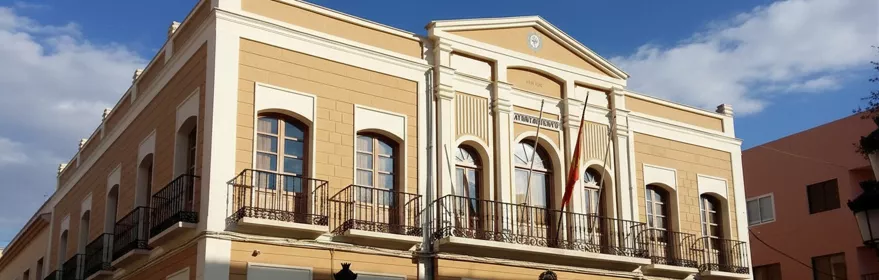Vista en ángulo de un edificio consistorial de estilo neoclásico, con fachada de color beige y blanco y balcones con rejas de hierro forjado, bajo un cielo con nubes.