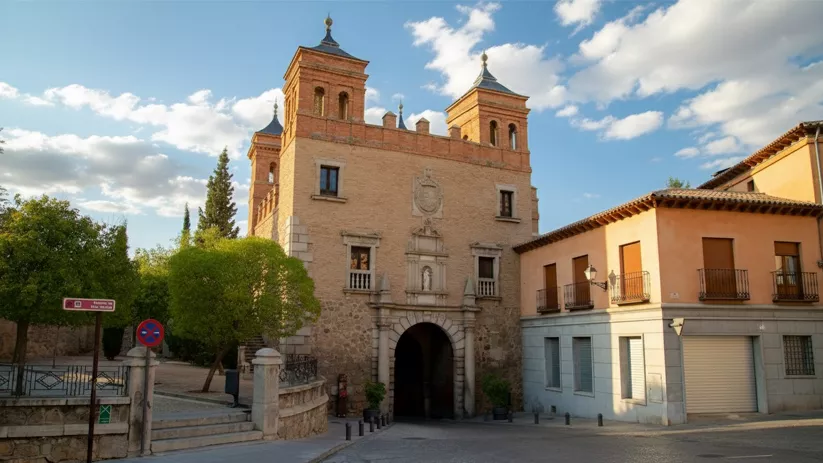 Vista frontal de una puerta de ciudad histórica, con una torre central con dos campanarios y una gran arcada de piedra, flanqueada por edificios residenciales.