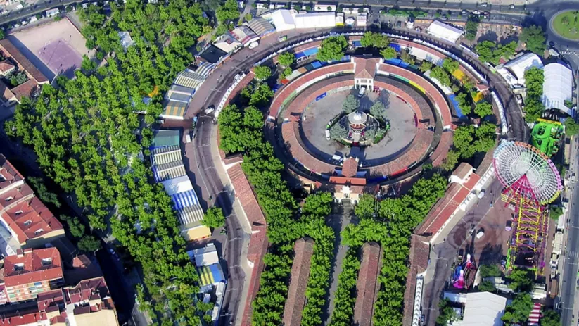 Vista aérea diurna de un recinto ferial circular rodeado de arbolado, con templete central, puestos y atracciones en el perímetro.