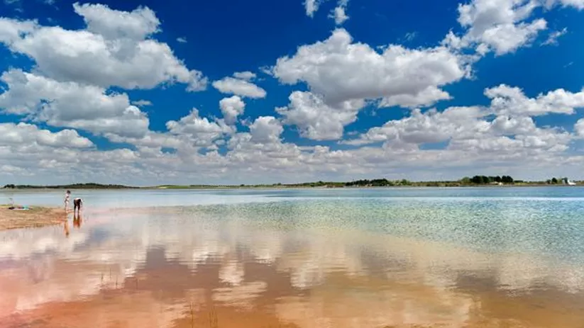 Orilla de laguna con agua poco profunda y cielo con nubes reflejadas.