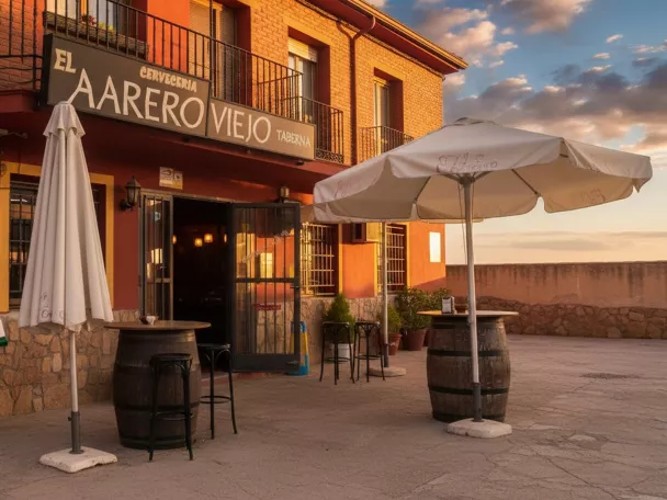 Entrada de taberna con sombrillas, barriles y fachada rojiza al atardecer.