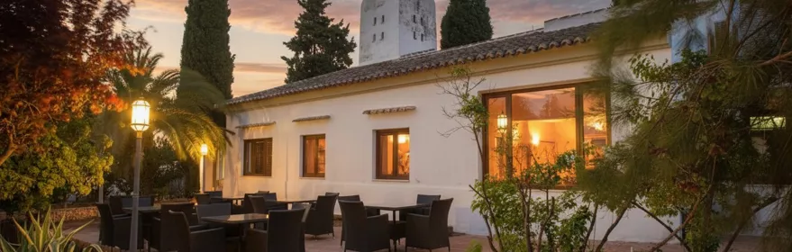 Terraza exterior con mesas y sillas junto a un edificio iluminado al atardecer.