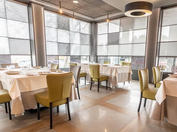 Comedor luminoso con mesas vestidas, sillas tapizadas y grandes ventanales con estores.