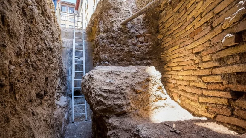 Zanja arqueológica estrecha con muros de ladrillo y escalera metálica.