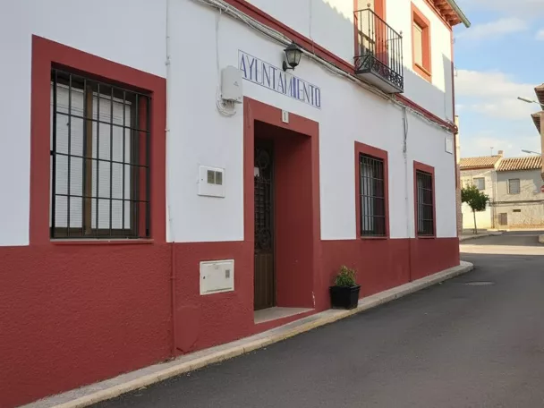 Una calle de pueblo con edificios residenciales blancos con bases rojas y ventanas con rejas de hierro, y un edificio de administración local con un cartel que dice "Ayuntamiento".