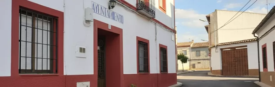 Una calle de pueblo con edificios residenciales blancos con bases rojas y ventanas con rejas de hierro, y un edificio de administración local con un cartel que dice "Ayuntamiento".