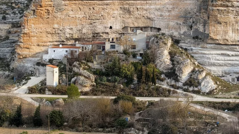 Casas integradas en un acantilado de roca, con huertos y camino en primer plano.