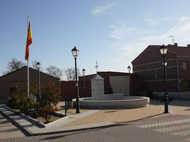 Vista panorámica de una plaza de pueblo con una fuente central de piedra, farolas de estilo antiguo, una bandera nacional en un mástil y edificios residenciales de ladrillo al fondo bajo un cielo despejado.