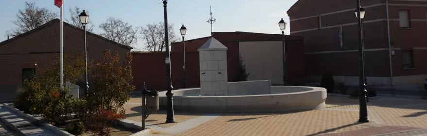 Vista panorámica de una plaza de pueblo con una fuente central de piedra, farolas de estilo antiguo, una bandera nacional en un mástil y edificios residenciales de ladrillo al fondo bajo un cielo despejado.