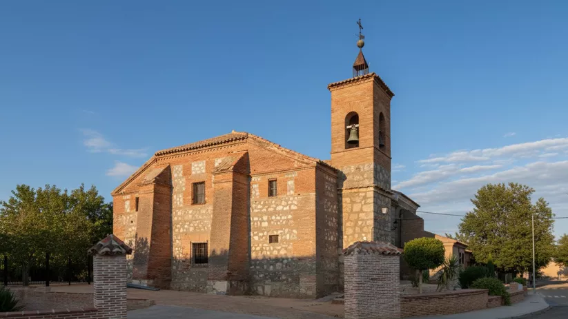 Perspectiva de una iglesia construida con una mezcla de piedra y ladrillo, con una torre campanario cuadrada coronada por una veleta, rodeada por un muro bajo y vegetación.