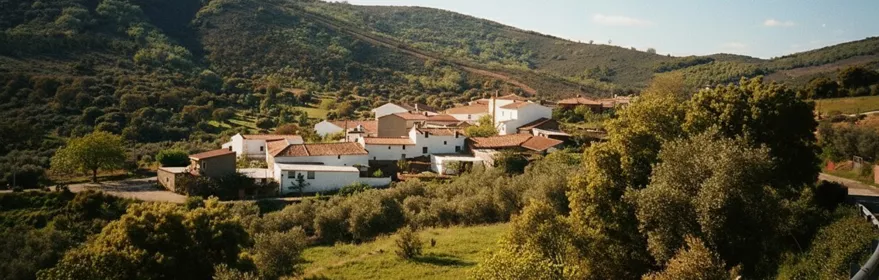 Vista panorámica de un pequeño pueblo de casas blancas con tejados de teja roja, enclavado en un valle verde rodeado de colinas cubiertas de vegetación bajo un cielo azul despejado.