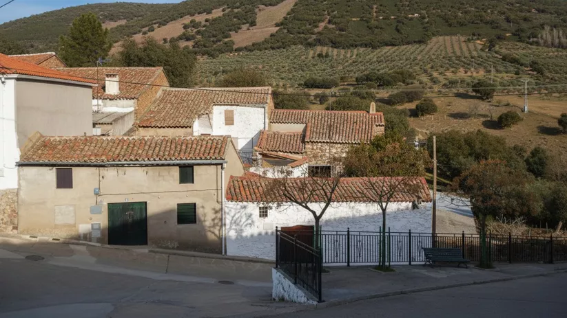 Perspectiva lateral de una casa rural de paredes blancas y tejado de teja roja, situada en una ladera con una valla negra en primer plano. Detrás de la casa, se extiende una colina verde cubierta de olivos y otra colina más alta con vegetación.