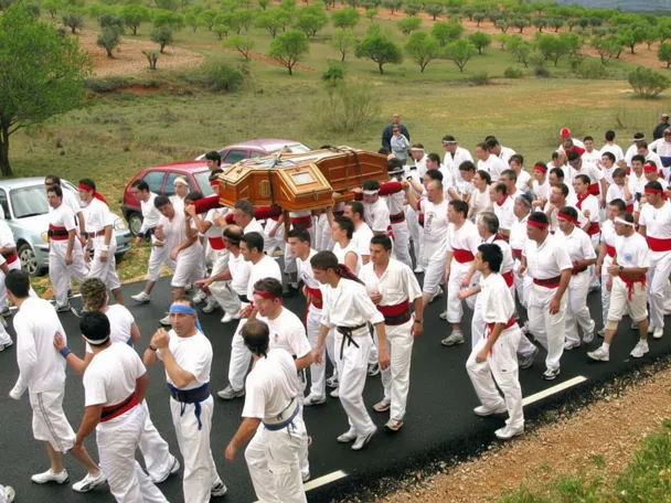 Gran grupo de participantes vestidos de blanco portando un féretro en una carretera junto a coches.