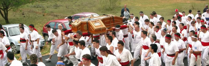 Gran grupo de participantes vestidos de blanco portando un féretro en una carretera junto a coches.