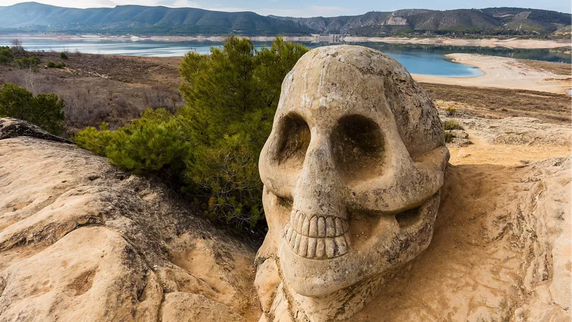 Escultura de calavera tallada en roca junto a un embalse.