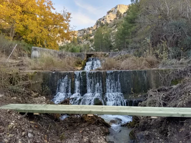 Pequeña cascada escalonada en un canal, con pasarela de madera y montañas al fondo.