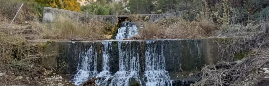 Pequeña cascada escalonada en un canal, con pasarela de madera y montañas al fondo.