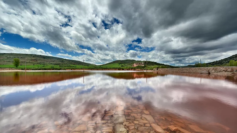 Balsa rojiza con reflejo de nubes y colinas verdes.